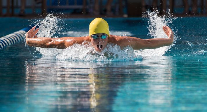 Male swimmer swimming in a lap pool wearing a yellow swim cap and goggles with visible arms and water splashing. 